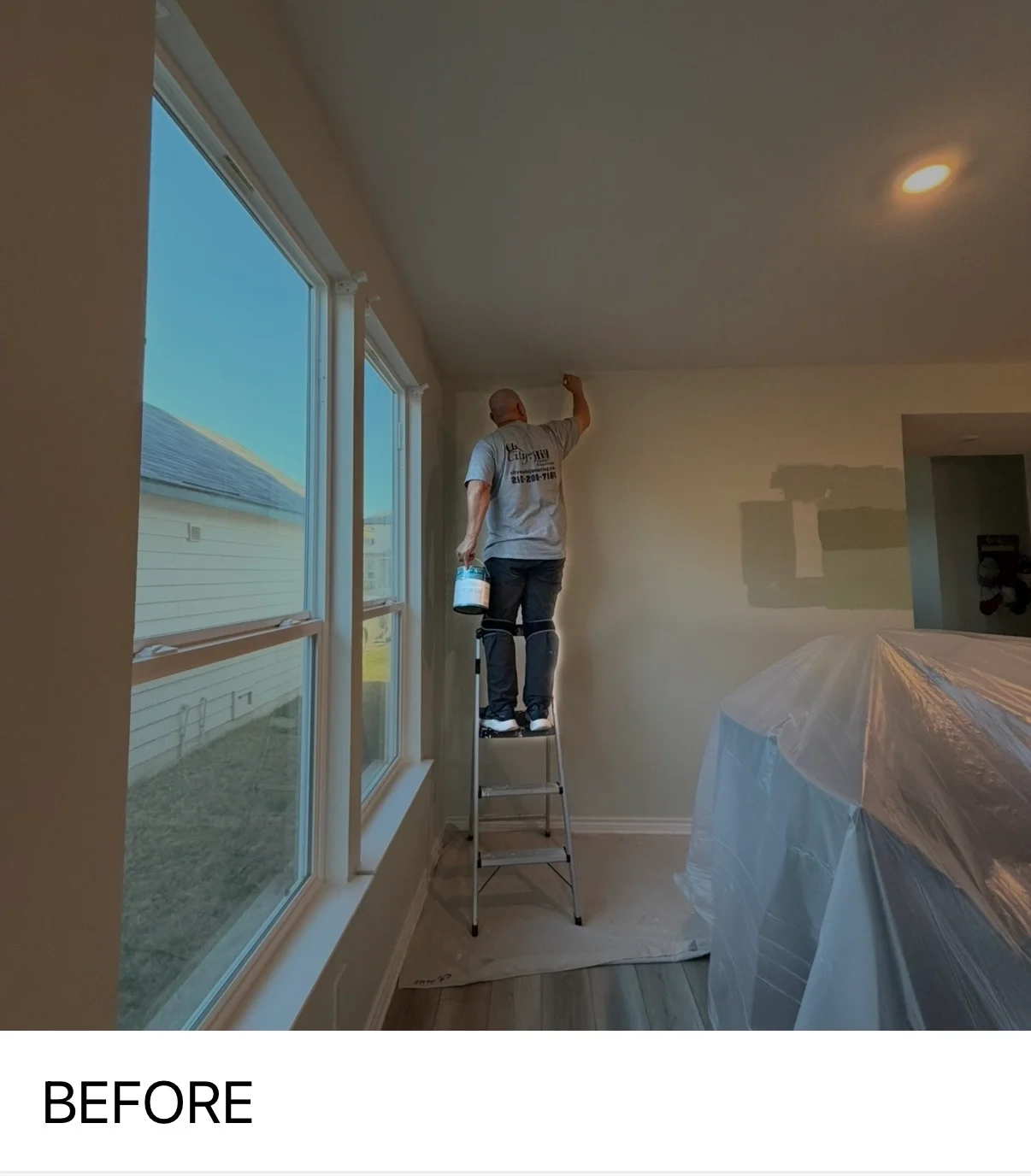 Painter on ladder applying primer to interior walls near large windows in residential home