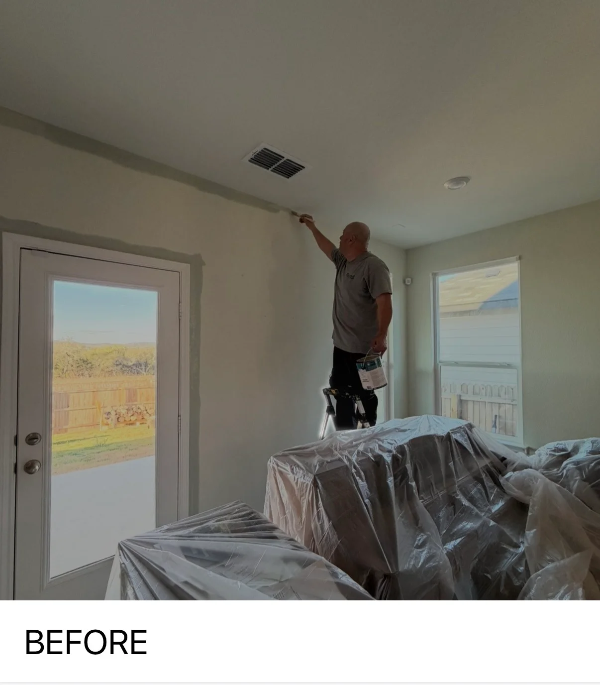 Painter applying finish coat to ceiling in bright room with protected furniture and scenic landscape views
